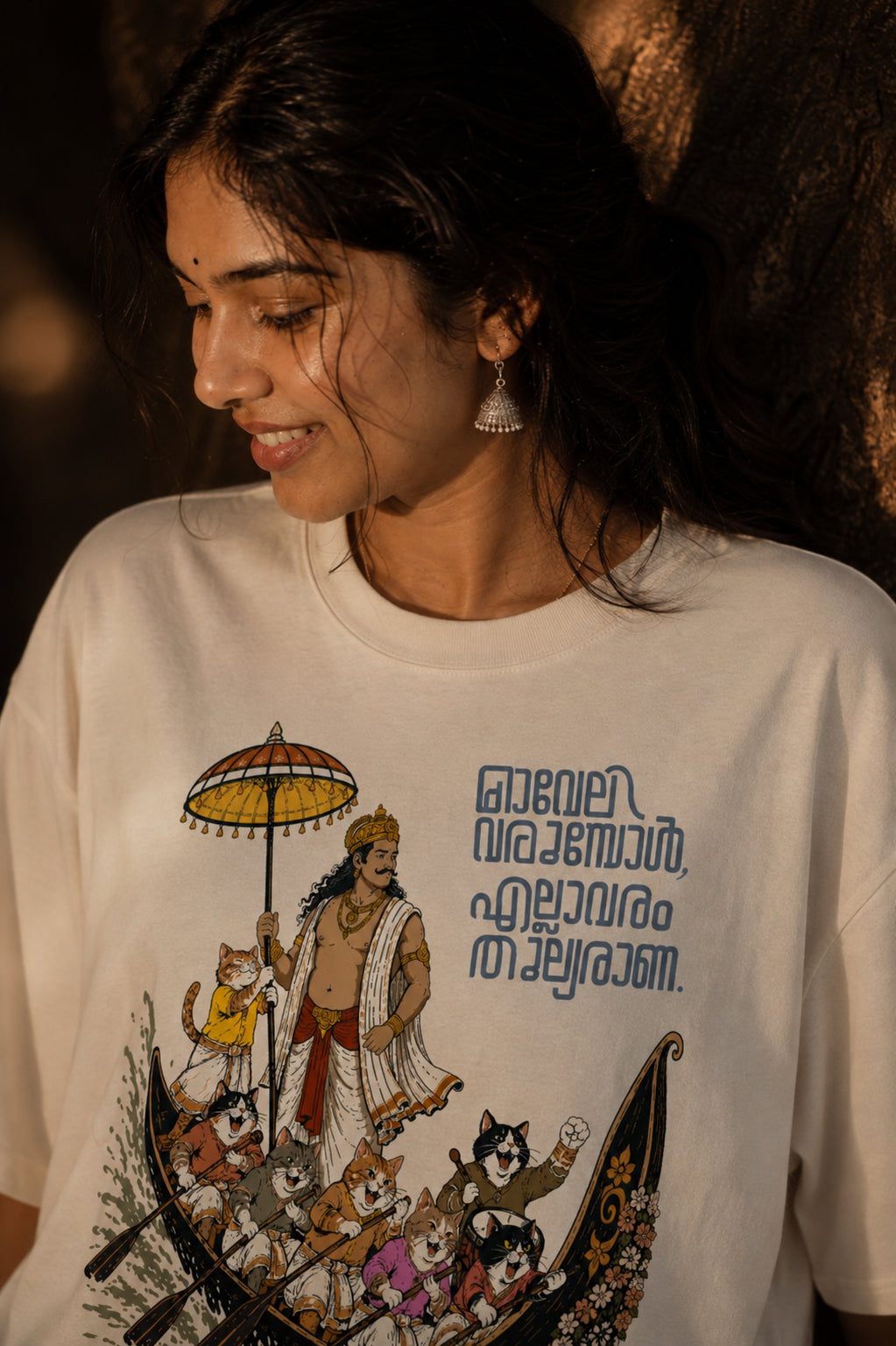 Woman wearing Mild Popie's All Are Equal oversized graphic T-shirt featuring Mahabali, Kerala's iconic snake boat race, cats, Indian dogs, and Malayalam typography inspired by Onam.