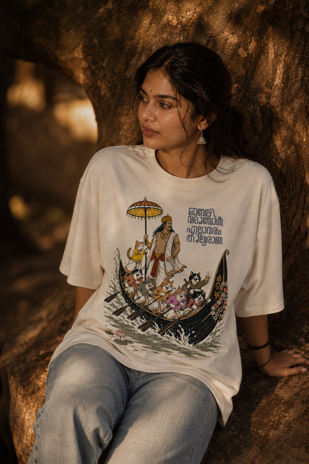 Woman wearing Mild Popie's All Are Equal oversized graphic T-shirt featuring Mahabali leading a Kerala snake boat race with cats and Indian dogs during Onam.