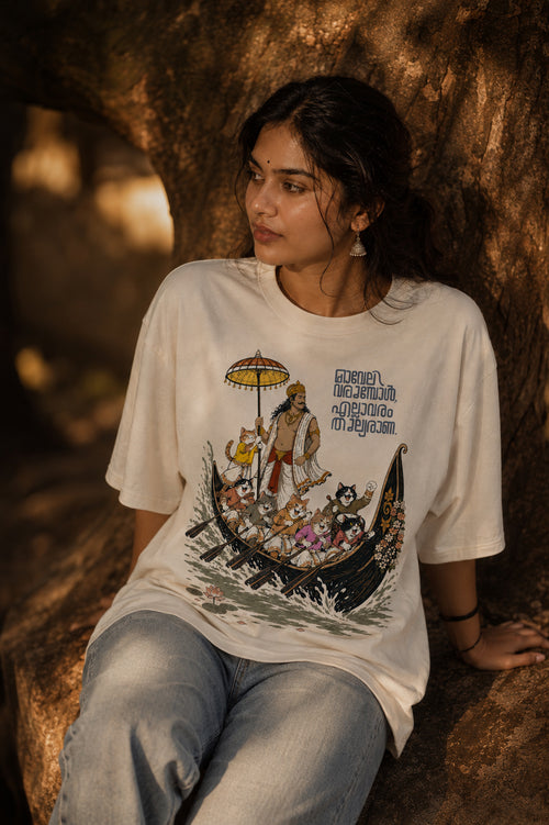 Woman wearing Mild Popie's All Are Equal oversized graphic T-shirt featuring Mahabali leading a Kerala snake boat race with cats and Indian dogs during Onam.